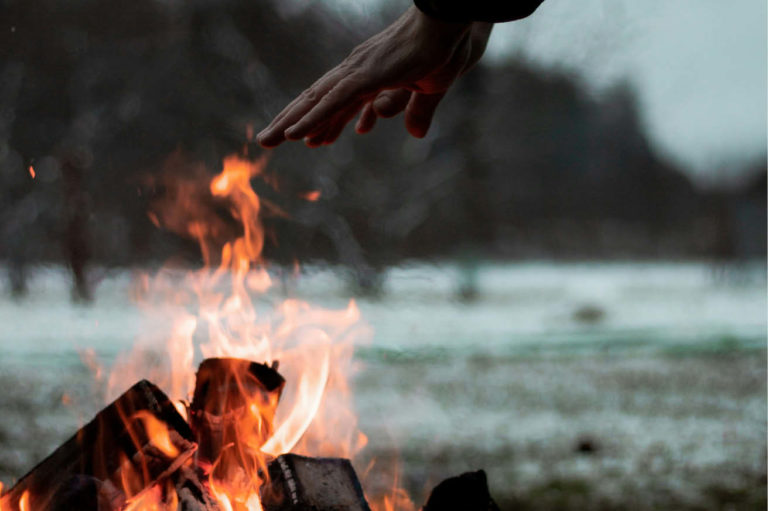 Hender som varmer seg på et bål i skumringen. Det er vinter og snø på bakken.