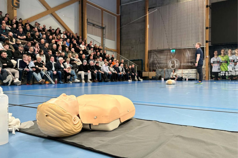 Mange mennesker sitter på tibunen og hører på en instruktør fra Blostrupmoen undervise i førstehjelp. i bilde ser du også en treningsdukke