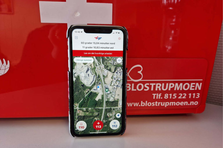 En telefon står forran en førstehjelpskoffert fra Blostrupmoen. Den er slått opp på appen hjelp 113, hvor den søker etter nærmeste hjertestarter